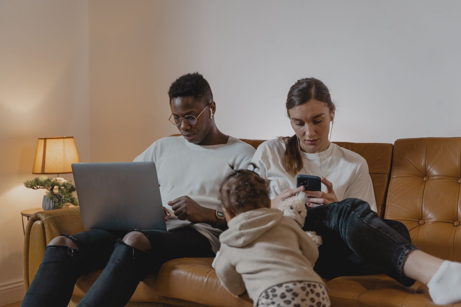 A family sitting on a couch using a laptop and phone while a toddler plays nearby. Cozy indoor setting.